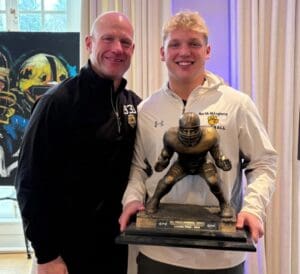 Two people smiling, one holding a football trophy. North Allegheny logo visible on jacket.