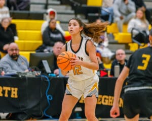 Basketball player in white jersey dribbling at a game, spectators in the background watching the match.