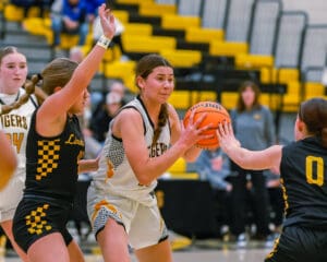 Girls' high school basketball game, player in white holds ball while being defended by two players in black uniforms.