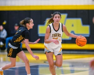 High school girls basketball game action, player in white jersey dribbling past defender on court.