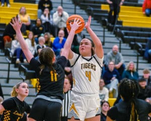 Basketball player in white jersey shoots over defenders in black during a game, with spectators in the background.