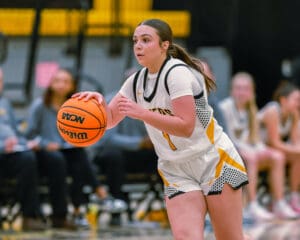 A basketball player dribbles on the court in an intense college game, wearing a white and yellow jersey.