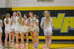 Girls' basketball team huddles on court in white jerseys, preparing for a game.