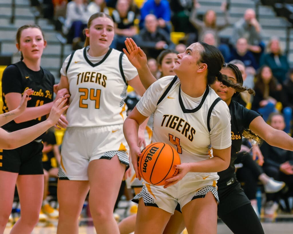 High school basketball game: player in white uniform holding ball, surrounded by opponents, aiming for the basket.