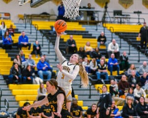 High school basketball player jumps for a layup shot in a game, with spectators in the background.