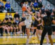 Female basketball player in white jersey jumps to shoot during a game against team in black and yellow uniforms.