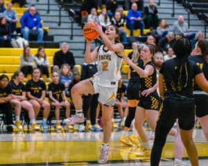 Female basketball player in white jersey jumps to shoot during a game against team in black and yellow uniforms.