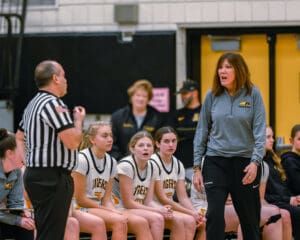 Female basketball coach engaged in discussion with referee, players on bench observing, during a high school game.