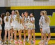 High school girls' basketball team in white uniforms huddling on the court.