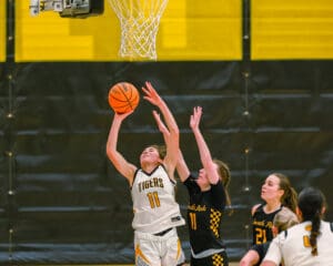 Girls' basketball game action with player jumping to score near the hoop, opponents attempting to block.