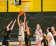Girls' basketball game action with player jumping to make a basket, surrounded by competing teammates and opponents.
