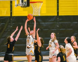 Girls' basketball game action with player jumping to make a basket, surrounded by competing teammates and opponents.