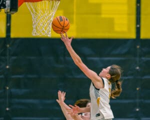 Basketball player in white jersey making a layup during a game, reaching for the hoop with orange ball.