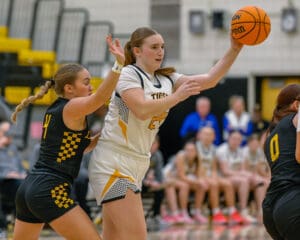 Girls' basketball game action, player in white passing under defensive pressure, spectators visible in the background.