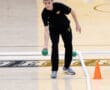 Person playing bocce ball indoors, focusing on aiming with green ball on a wooden court.