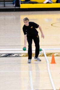 Person playing bocce ball indoors, focusing on aiming with green ball on a wooden court.