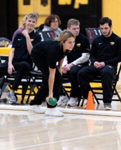 Person lining up a bocce ball shot indoors, teammates watching attentively on the sidelines.
