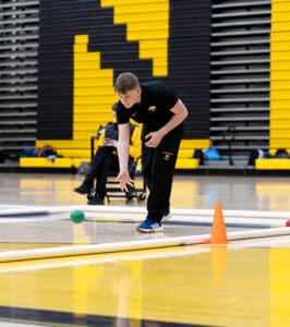Person playing bocce in a gym, aiming at a green ball near an orange cone on the court.