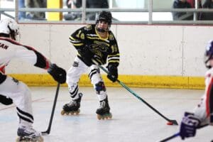 Youth roller hockey player in action, wearing black and yellow gear, competes for the puck on an indoor rink.