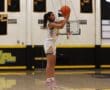 Basketball player in mid-air shot during a game, wearing a white and yellow uniform on a gym court.
