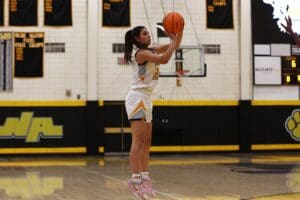 Basketball player in mid-air shot during a game, wearing a white and yellow uniform on a gym court.