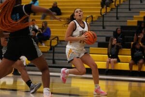 Basketball player driving to the hoop during a game, with opposing defender in pursuit on a brightly lit court.