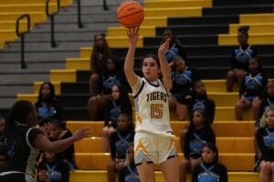 Basketball player in Tigers uniform shoots ball during game, with cheerleaders in the background on bleachers.