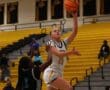 Basketball player in white jersey makes a layup during a game on a yellow bleacher court.