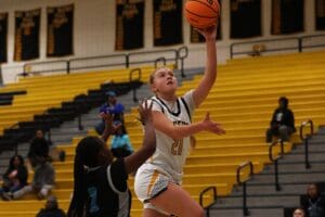Basketball player in white jersey makes a layup during a game on a yellow bleacher court.
