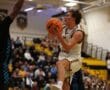 Basketball player in white uniform jumps for a shot during an intense game in a crowded gymnasium.