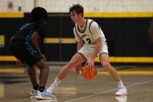 Basketball player dribbles during a high school game, wearing a white uniform, facing a defender on court.