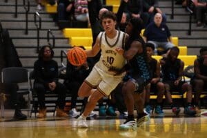 Basketball players compete on the court, one in white jersey dribbling while guarded by opponent in black.