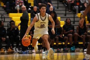 Basketball player dribbles down court during a high school game, with focused teammates and spectators watching.