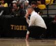 Basketball coach deep in thought during a game, squatting beside the court near a table labeled Tigers.