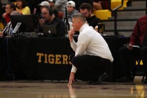 Basketball coach deep in thought during a game, squatting beside the court near a table labeled Tigers.
