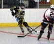 Roller hockey players competing on an indoor rink, one in black and yellow, the other in white and red, with sticks engaged.