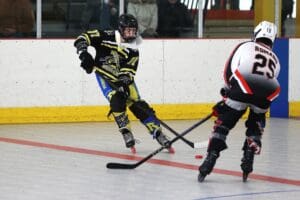 Roller hockey players competing on an indoor rink, one in black and yellow, the other in white and red, with sticks engaged.