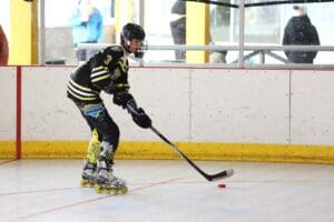Roller hockey player in action on indoor rink, wearing black and yellow gear, ready to hit puck with stick.