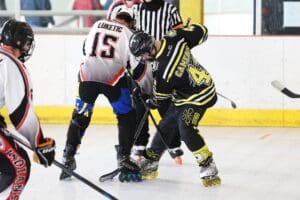 Roller hockey faceoff between players in black and white jerseys on an indoor rink, overseen by a referee.