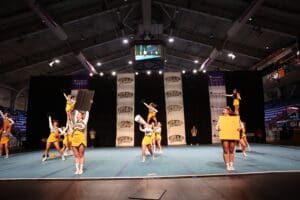 Cheerleaders performing stunts at indoor competition, yellow uniforms, large banners in the background.