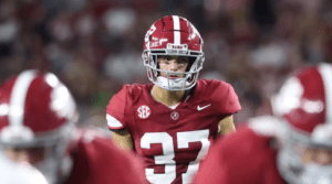 Football player in red Alabama jersey during a game, wearing a red helmet with a focused expression.