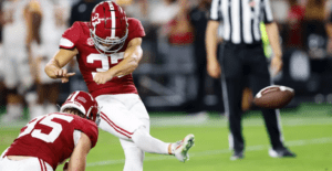Football player in red jersey kicking a field goal during a game, with referee in the background.