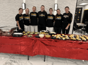 Women's team in Tigers shirts standing behind a buffet table with salad and bread.