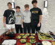 Four young people standing behind a table with salad bowls, wearing casual outfits and smiling.