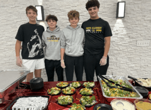 Four young people standing behind a table with salad bowls, wearing casual outfits and smiling.