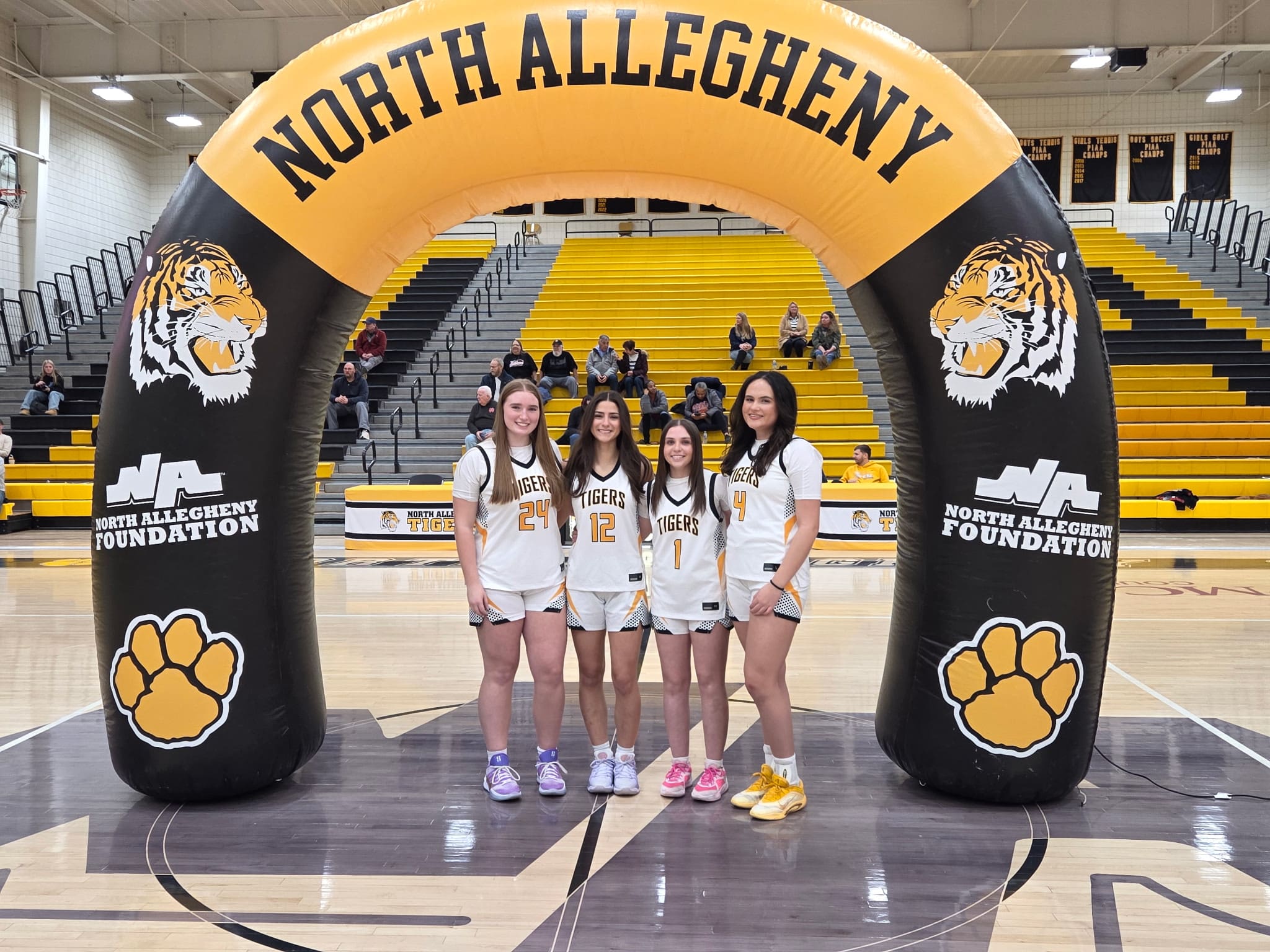Four female basketball players in North Allegheny Tigers jerseys stand under an inflatable arch in a gym.