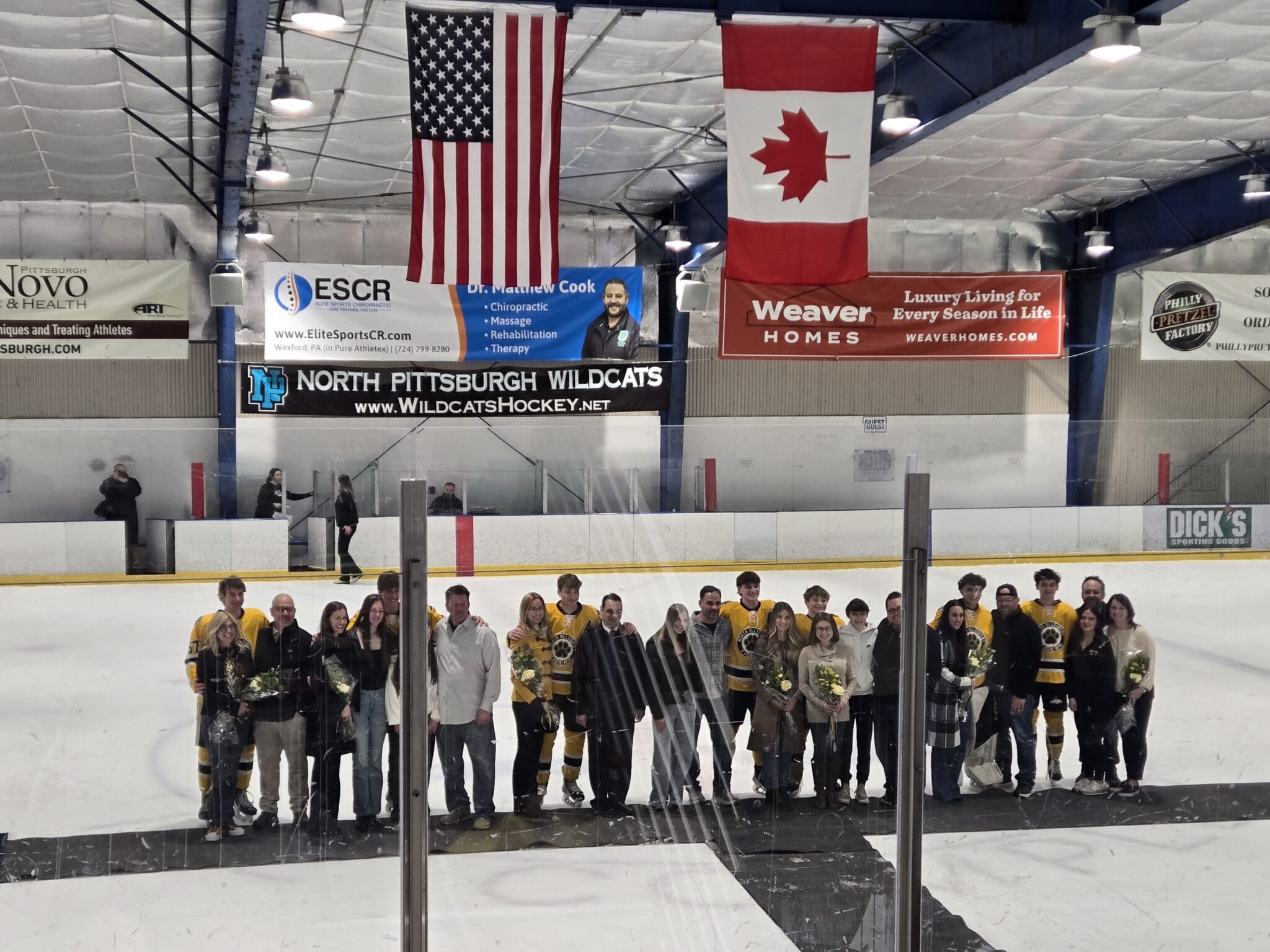 Hockey players and families pose on ice rink at North Pittsburgh Wildcats event under flags and banners.