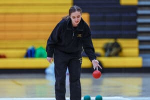 Person playing bocce indoors, wearing a black tracksuit, holding a red bocce ball with yellow bleachers in the background.