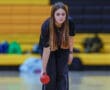 Young bowler focuses intently while holding a red bowling ball, preparing to roll it down the lane in a gym setting.