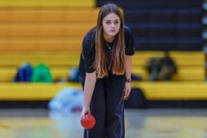 Young bowler focuses intently while holding a red bowling ball, preparing to roll it down the lane in a gym setting.